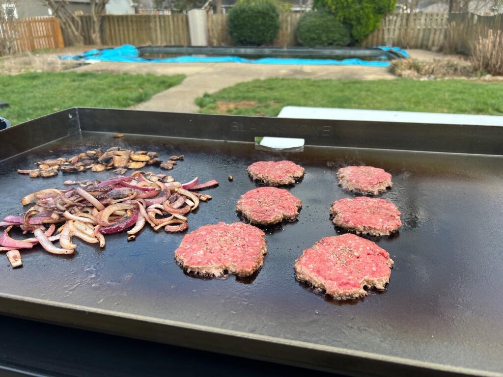 Ground beef balls smashed flat into patties on the griddle, next to grilled onions