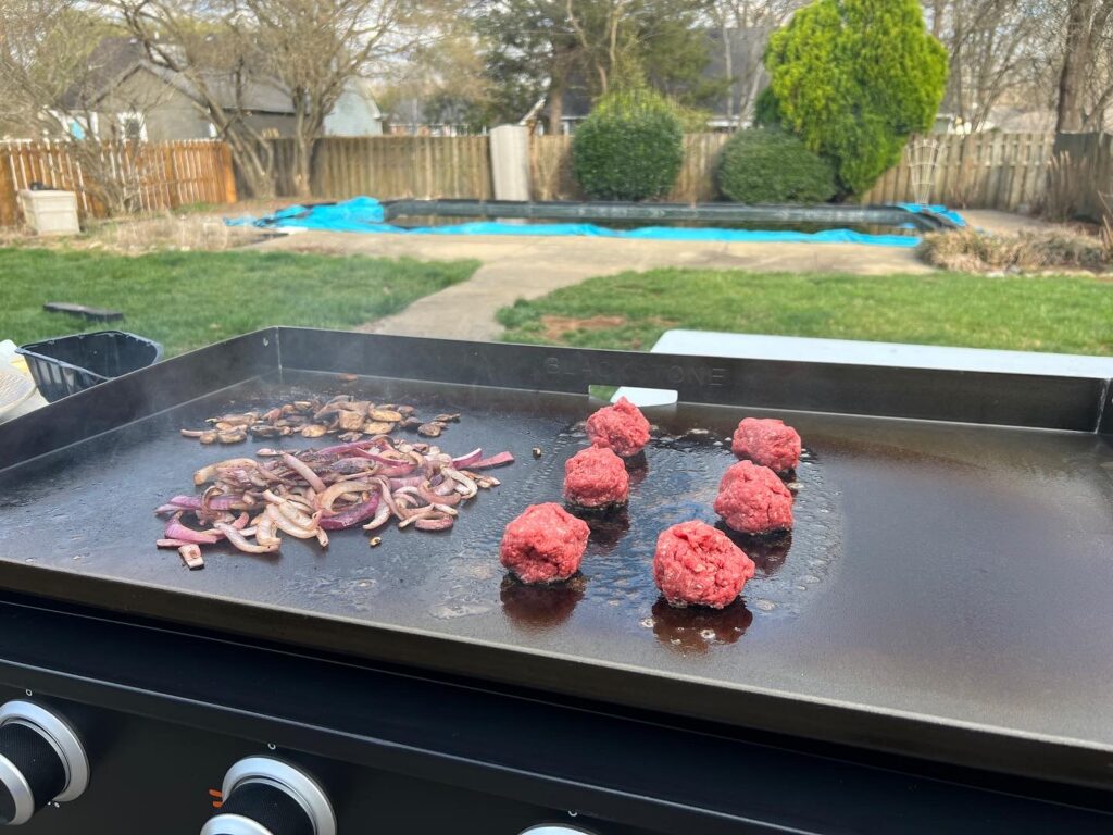 Raw ground beef balls on the Blackstone griddle next to grilled onions, before smashing