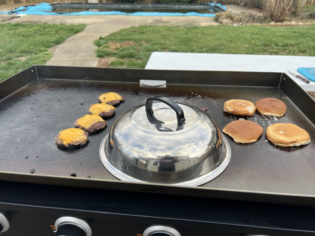 Smash burgers melting cheese under a metal dome, buns toasting alongside on the griddle