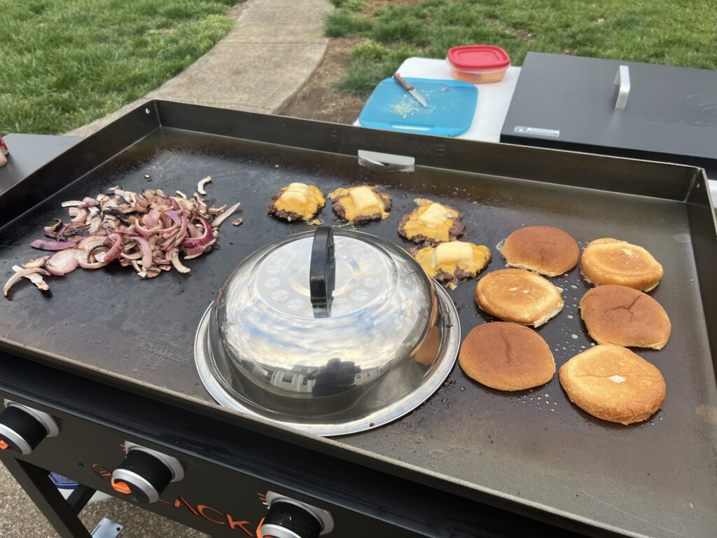 A second round of smash burgers melting cheese under the dome, with more buns toasting