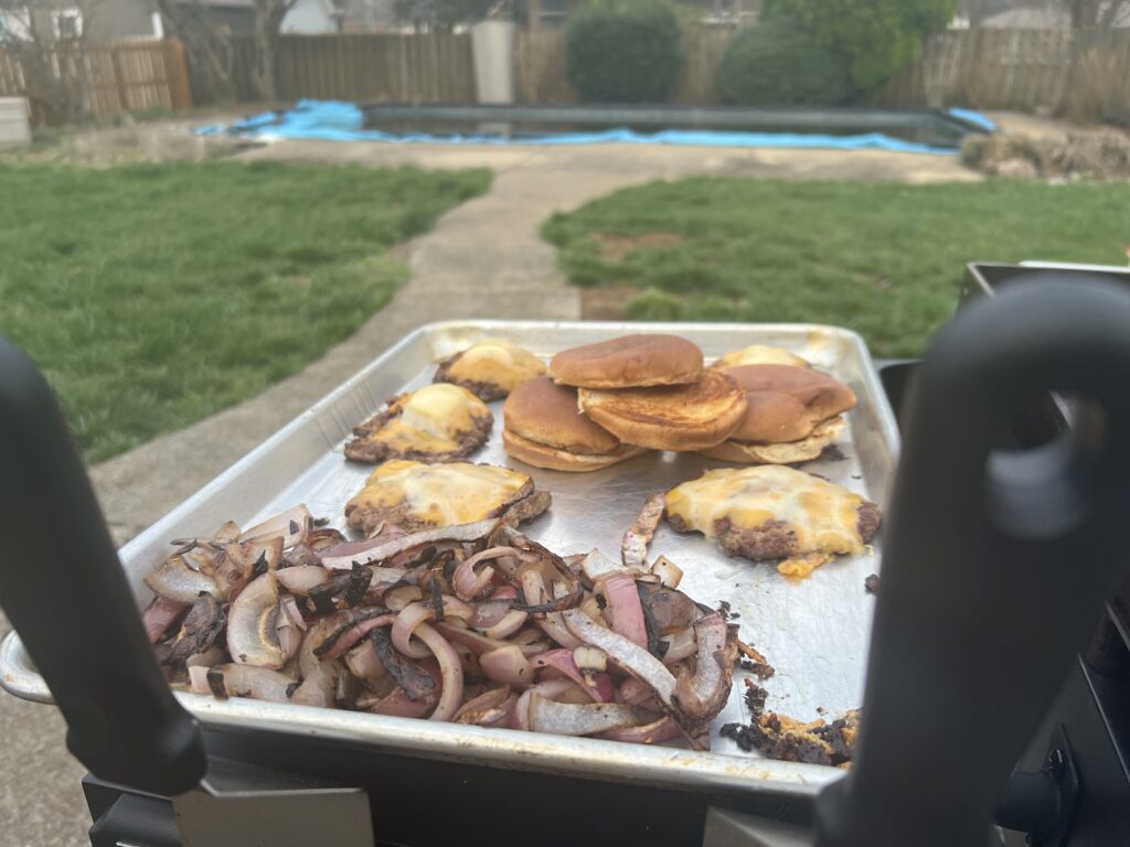 Finished smash burgers with melted cheese, grilled onions, and toasted buns, plated on a sheet pan