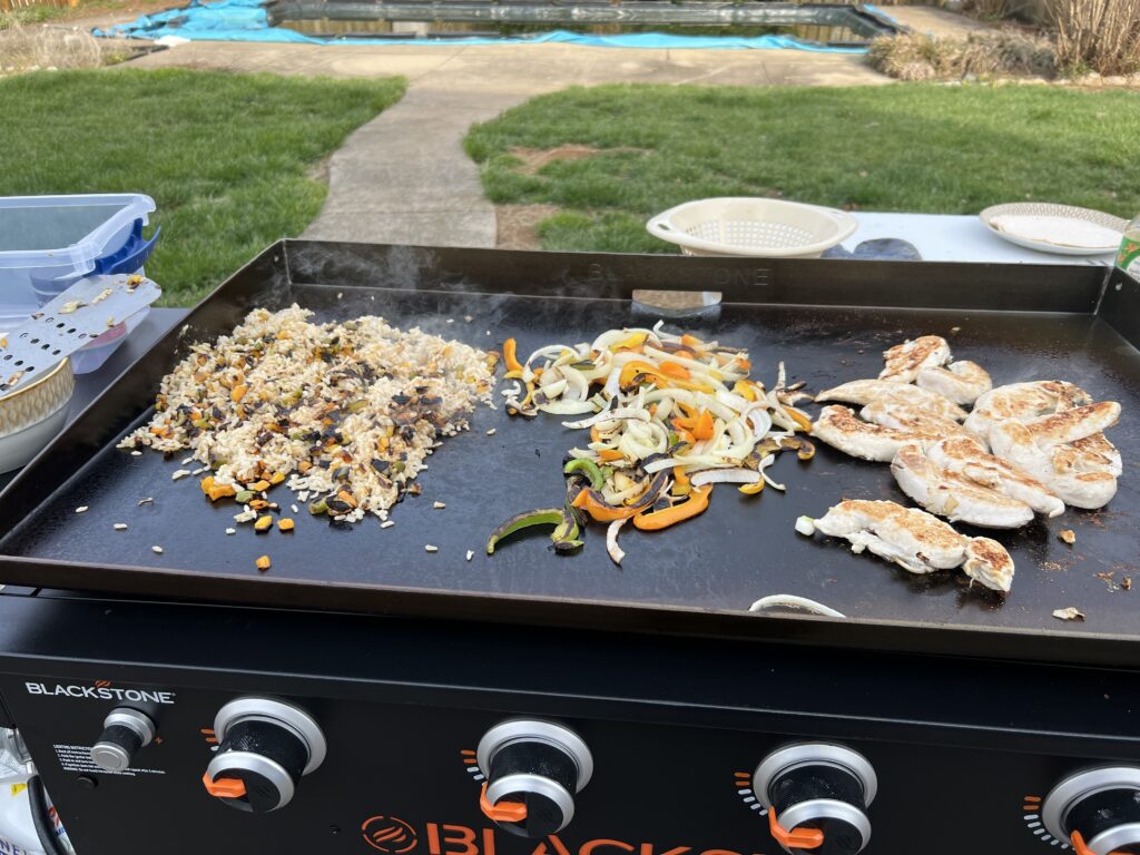 Rice, sliced peppers and onions, and grilled chicken cooking together on the griddle
