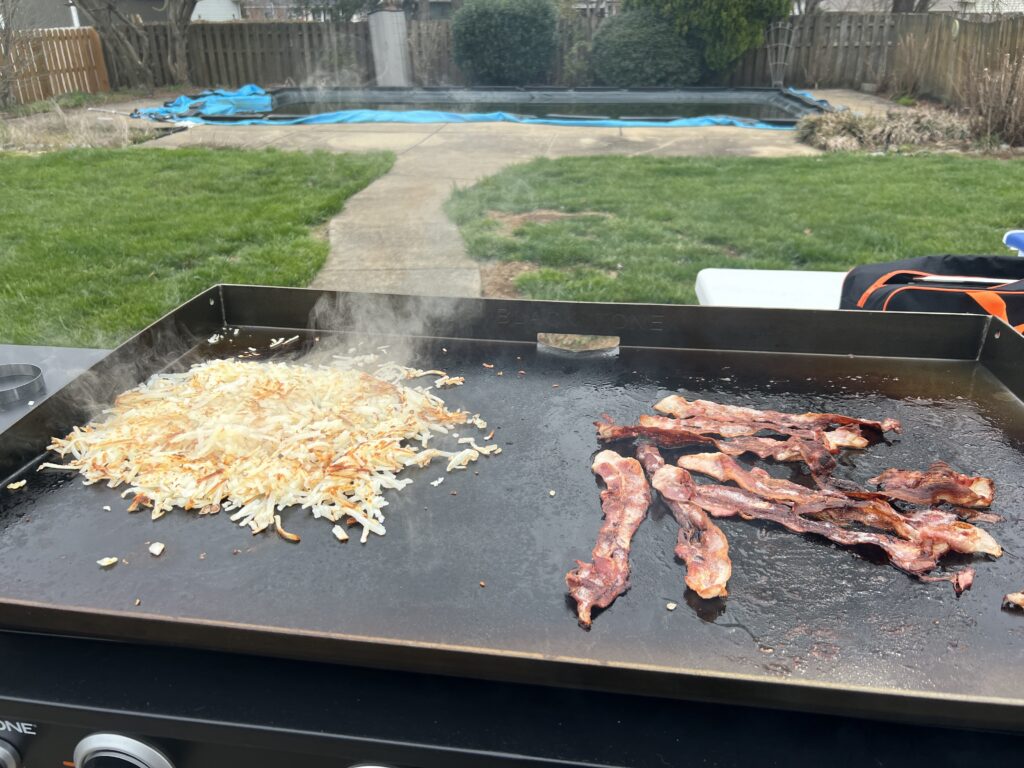 Bacon and hash browns cooking on the griddle, bacon starting to curl