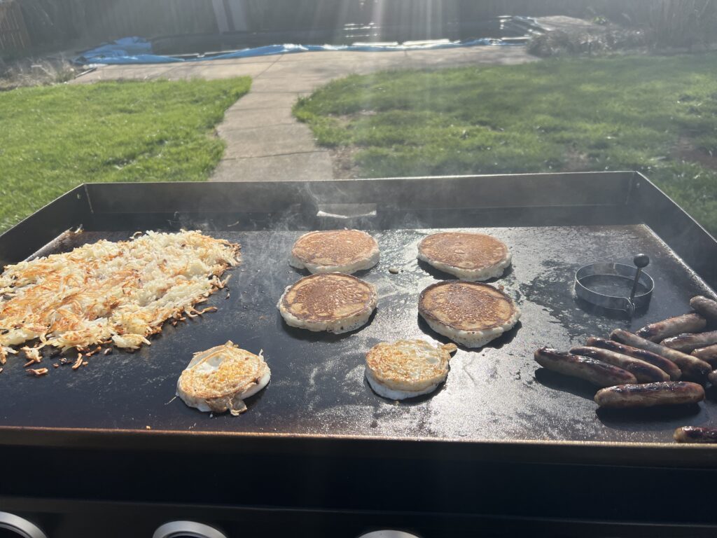 A better second breakfast attempt: hash browns, pancakes, and sausage links cooking together, with an egg ring on the griddle