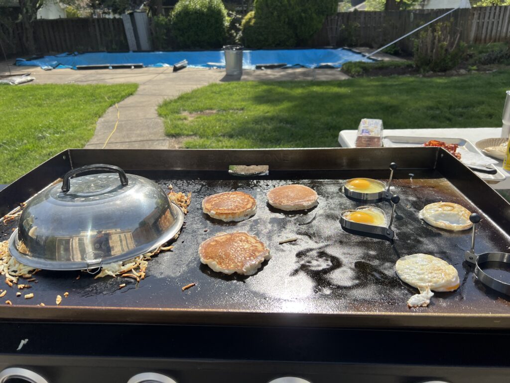 Eggs cooking in metal rings next to pancakes, with hash browns steaming under a dome