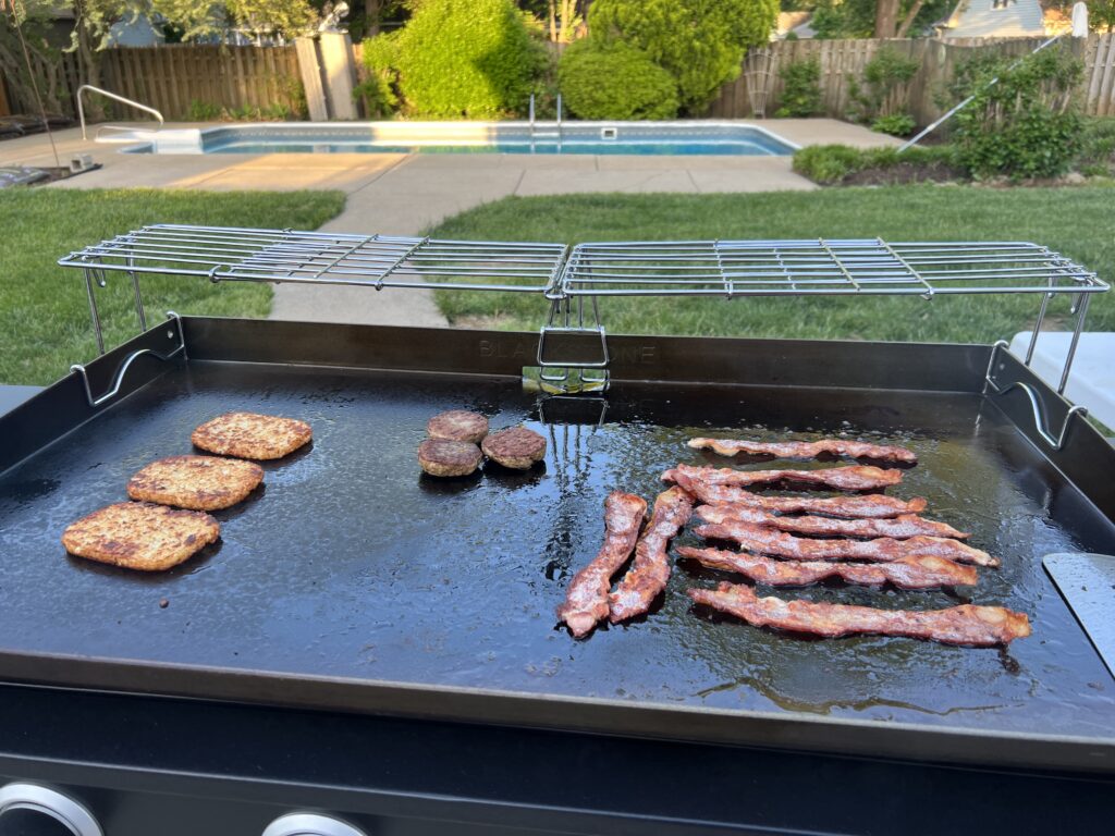 hash brown patties cooking next to sausage patties and bacon, with a warming rack overhead