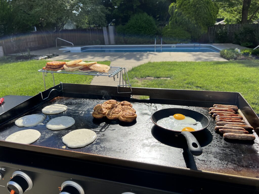 A full breakfast spread: cinnamon rolls, pancakes, eggs in a small skillet, and sausage links, with toast warming on a rack overhead