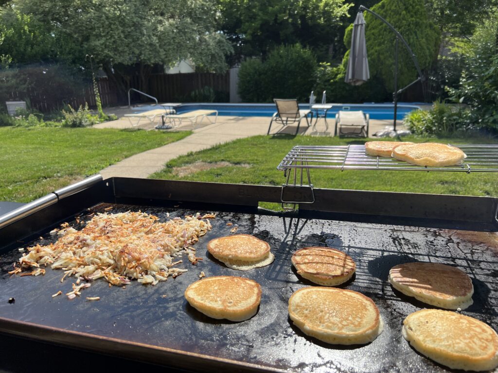 Hash browns and pancakes cooking on the griddle, with more pancakes and toast warming on the rack