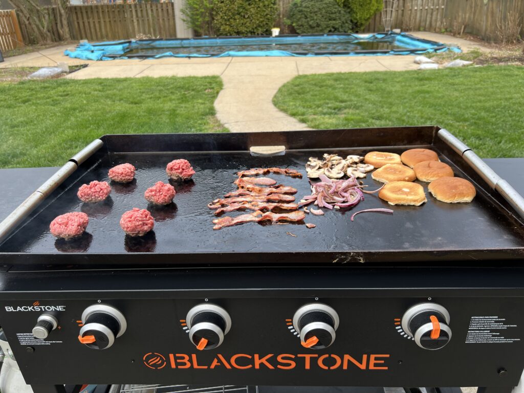 Beef balls, bacon, sliced onions, and mushrooms all cooking at once on the griddle, buns toasting alongside
