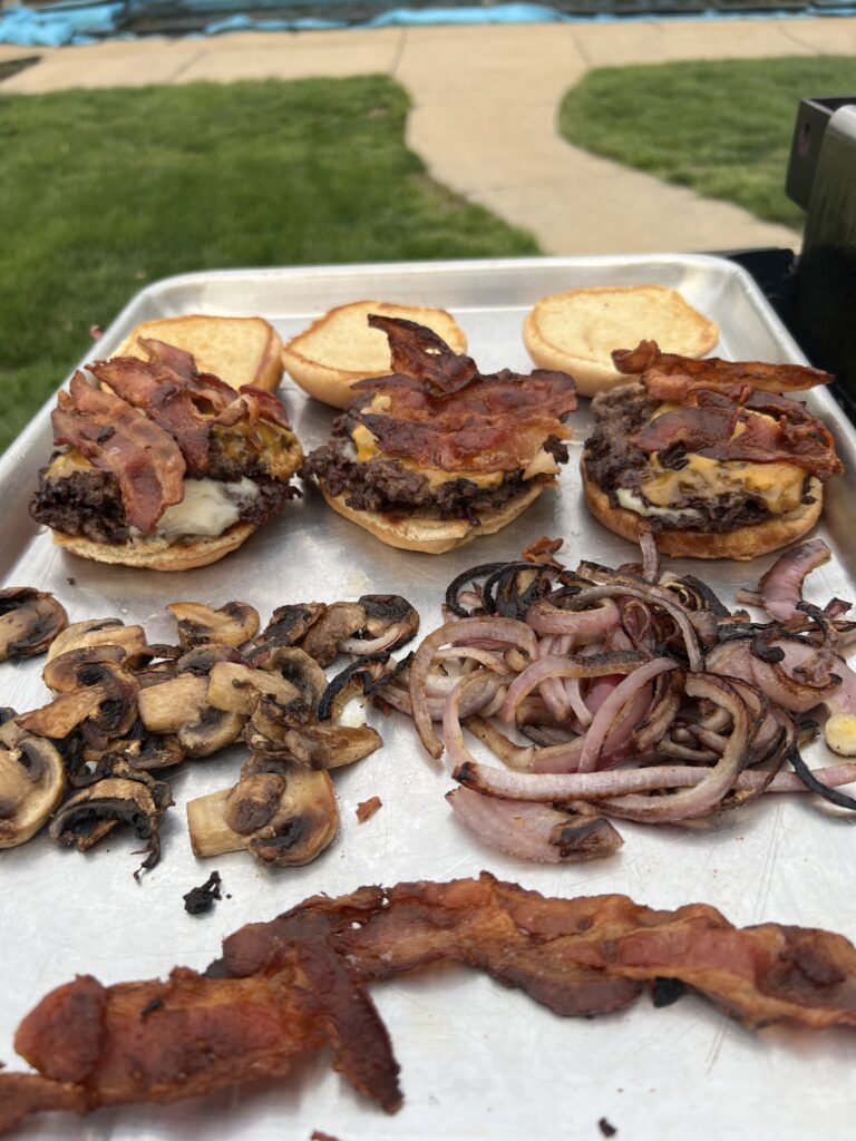 Finished beef smash burgers plated with grilled mushrooms, onions, and extra bacon on the side