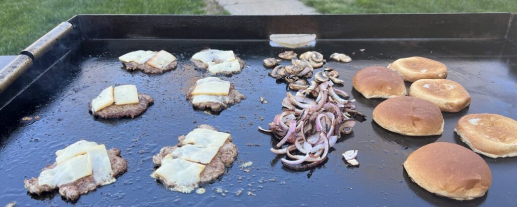 Pork smash burger patties with cheese melting, onions and mushrooms cooking alongside, buns toasting