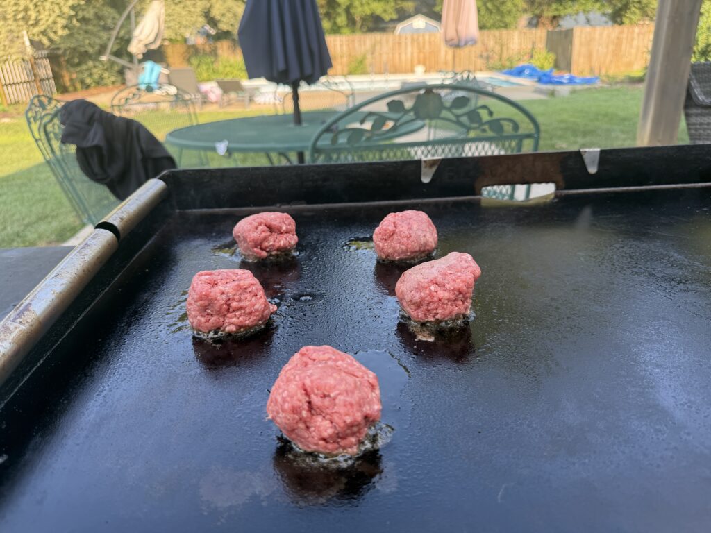 Five balls of ground beef searing in butter on a hot Blackstone griddle before being smashed