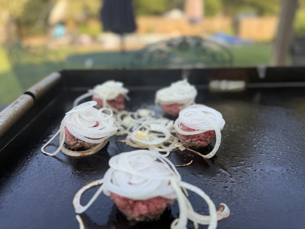 Thin sliced onions being smashed into ground beef patties on a Blackstone griddle for onion smash burgers