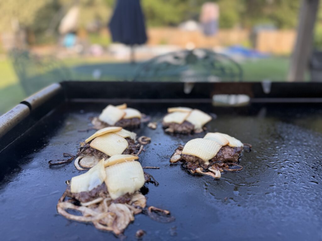 Flipped onion smash burger patties topped with slices of home-smoked white cheddar cheese melting on the Blackstone

